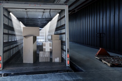 Several large, heavy retail merchandise cartons are stacked in an open delivery truck backed into a loading dock. Some of the cardboard cartons appear damaged. A manual fork lift hand truck holding a wooden pallet is waiting to help workers move the goods.