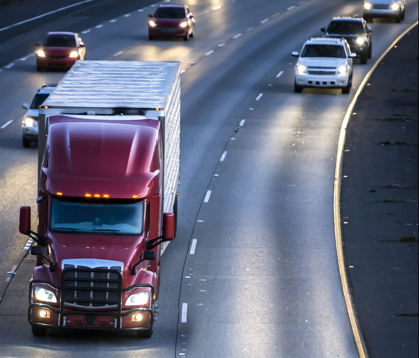 Dark red big rig semi truck transporting cargo in refrigerator semitrailer running on the wide multilines highway at night with turned on headlights