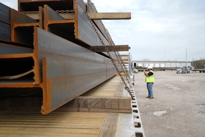 Steel beams loaded on a flatbed trailer with a truck driver in a hard hat and safety vest standing by truck