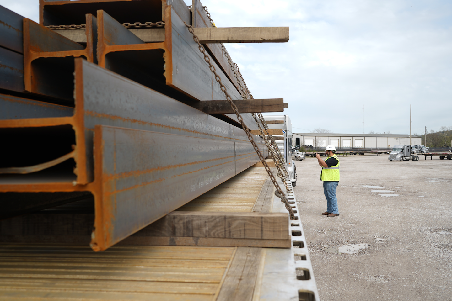 Steel beams loaded on a flatbed trailer with a truck driver in a hard hat and safety vest standing by truck