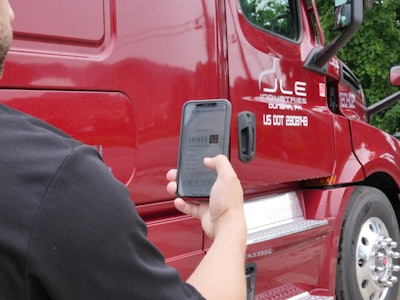 A person standing by a red semi-truck while holding a smart phone with DriverOS on screen