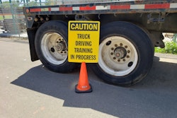 caution truck driving training in progress sign in front of the back wheels of a semi-truck