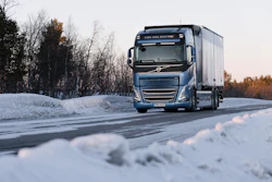 A blue truck going down a snowy road lined with trees.