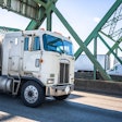Old fashioned cab over big rig semi truck transporting fuel in tank semi trailer running on the truss arched Interstate Bridge