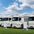 White electric trucks lined up on green grass, charging at electric stations
