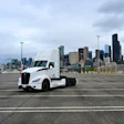 White Kenworth semi-truck parked in lot with Seattle skyline and Space Needle visible in background under cloudy sky