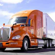 Orange Kenworth semi-truck with white trailer on parking lot against blue sky