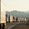 Long line of white semi-trucks on desert highway with mountain range in distance