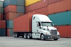 White semi-truck parked in front of stacked shipping containers in industrial yard