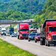 Red semi-trucks and passenger vehicles driving on a highway through green forested landscape
