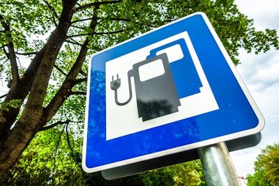 Blue road sign showing electric vehicle charging station symbol near trees