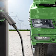 Green electric truck charging at station with wind turbines in background