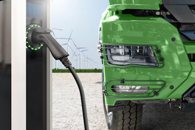 Green electric truck charging at station with wind turbines in background