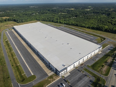 Aerial view of large white warehouse complex surrounded by forested landscape