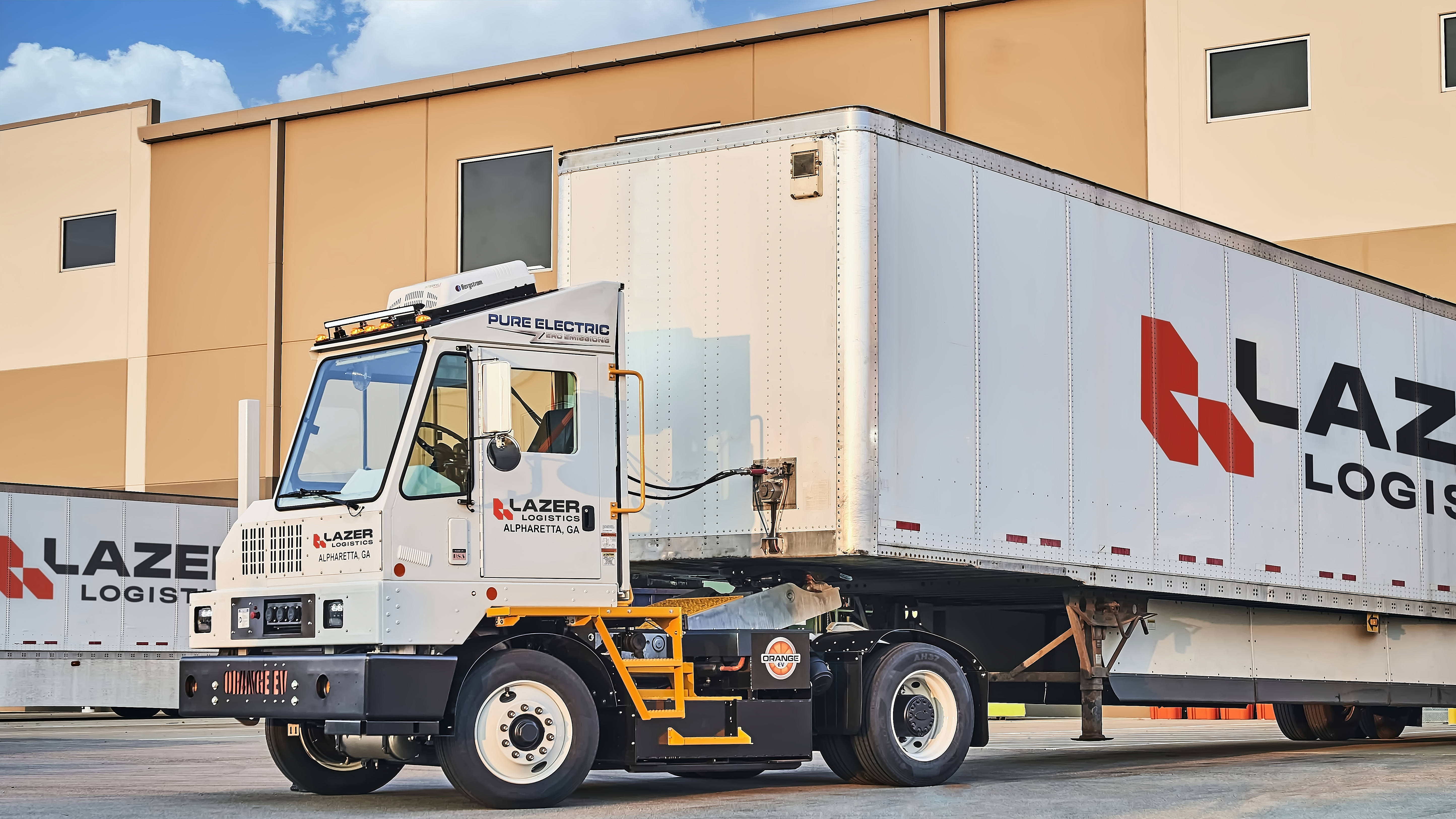An Orange EV yard tractor hauling a Lazer Logistics trailer at a loading dock.