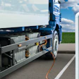 Blue electric truck charging at a white EV charging station with green field and blue sky background