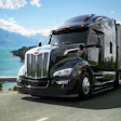 Black Peterbilt semi-truck driving along scenic mountain lake highway
