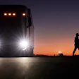 Large truck with bright headlights at sunset, person walking silhouetted against orange sky