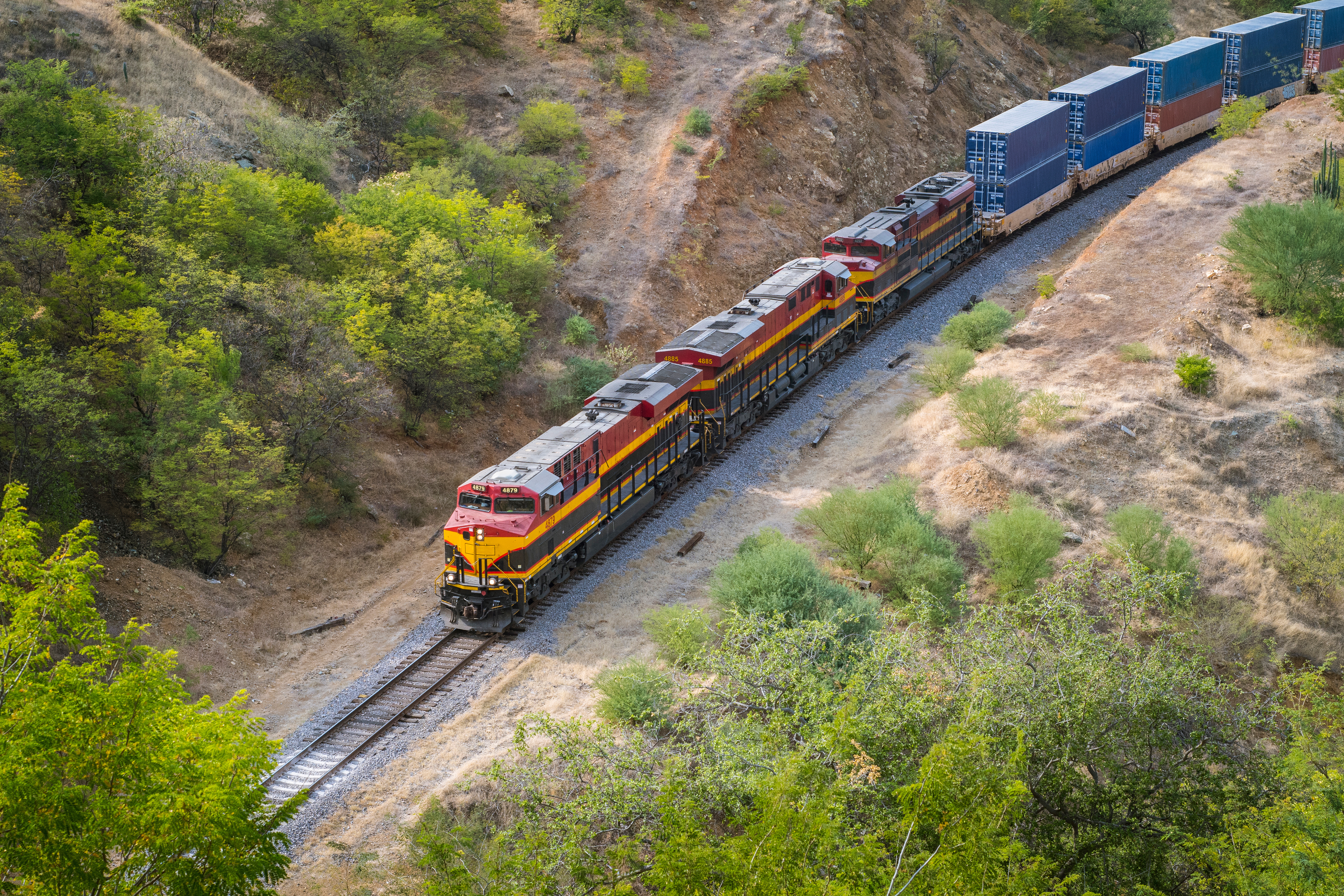 Freight train with red and yellow locomotives traveling through green mountainous terrain