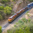 Freight train with red and yellow locomotives traveling through green mountainous terrain