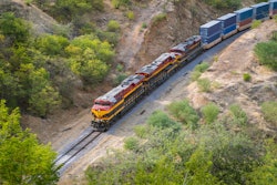 Freight train with red and yellow locomotives traveling through green mountainous terrain