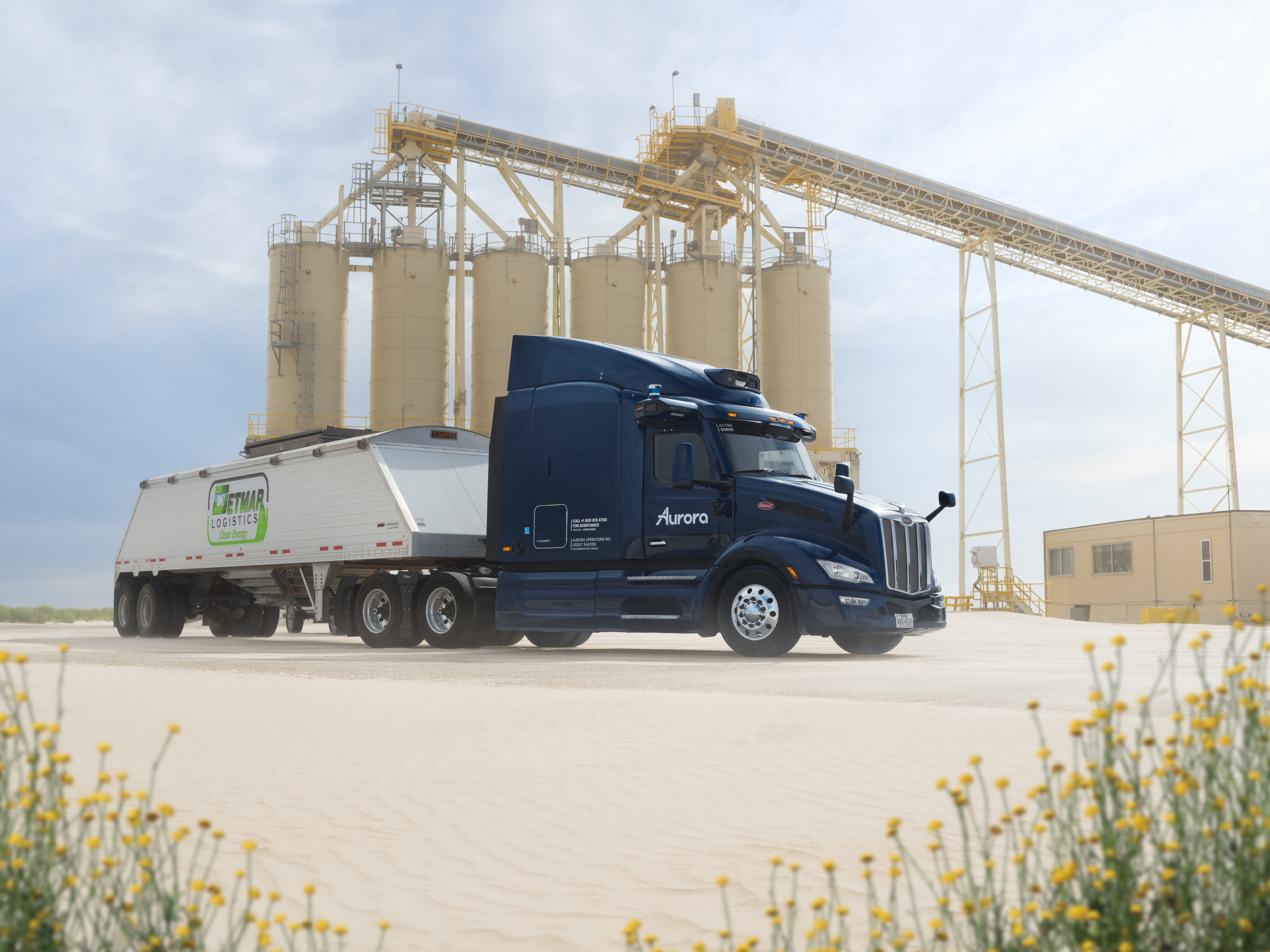 Dark blue Aurora semi-truck parked near industrial silos with yellow wildflowers