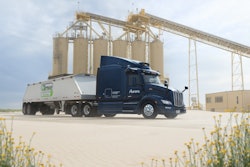 Dark blue Aurora semi-truck parked near industrial silos with yellow wildflowers
