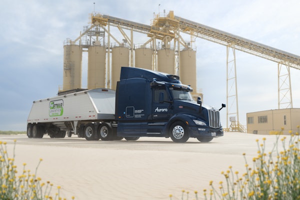 Dark blue Aurora semi-truck parked near industrial silos with yellow wildflowers