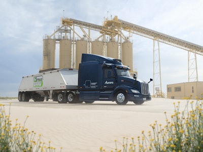 Dark blue Aurora semi-truck parked near industrial silos with yellow wildflowers