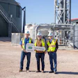 Three workers in safety vests at clean energy industrial facility with storage tanks