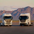 Two Nikola white semi-trucks parked on road with rugged mountain range in background