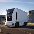 White autonomous semi-truck with sleek design parked at industrial facility with blue sky background