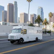 White XOS delivery truck driving on street with downtown Los Angeles skyscrapers in background