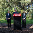 Three people in business suits hold a political protest sign in a wooded area with green trees in the background.