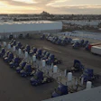 Aerial view of industrial logistics facility with blue trucks and trailers parked in organized rows beside a large warehouse building at dusk.