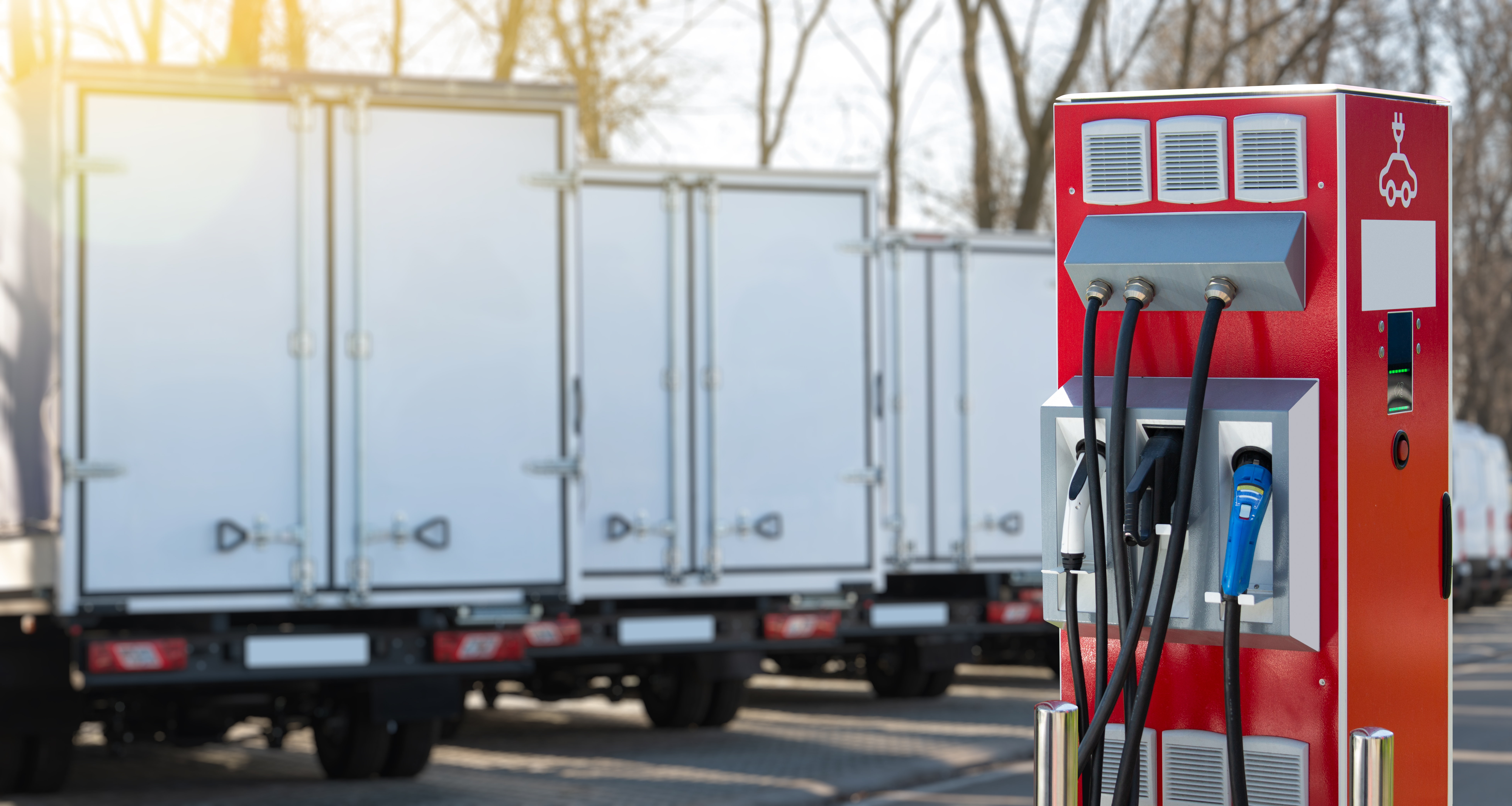 An electric vehicle charging station with multiple cables in an outdoor parking lot, with battery-electric-powered trucks in the background.