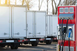 An electric vehicle charging station with multiple cables in an outdoor parking lot, with battery-electric-powered trucks in the background.