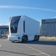 White autonomous electric truck with sleek cab design parked at industrial facility under clear blue sky