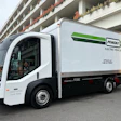 White Penske electric delivery truck with green branding parked in front of a modern residential building