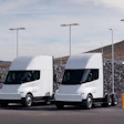 Two white Tesla Cybertrucks parked at an electric vehicle charging station with street lights and rocky hillside backdrop
