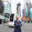 Professional man in navy suit posing before autonomous vehicle display in Times Square with skyscrapers and digital billboards