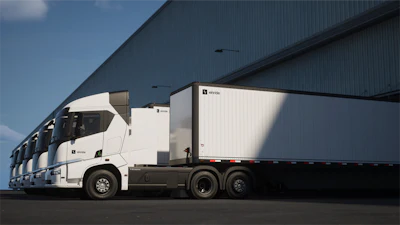 A lineup of Einride battery-electric semis with trailers attached parked at a warehouse loading dock.