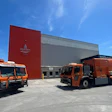 Orange waste management trucks parked outside an industrial facility with red and gray paneling under clear blue sky