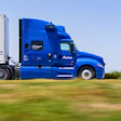 Blue Aurora semi-truck with white refrigerated trailer driving on highway through green fields