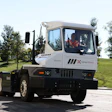 White and black Kalmar Ottawa electric terminal tractor with operator visible in cabin, parked on paved surface with green grass and trees in background