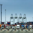 Busy container port terminal with stacked cargo containers, trucks, and gantry cranes under clear blue sky