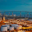 Industrial port facility with shipping containers, storage tanks, and bridge spanning the harbor at sunset