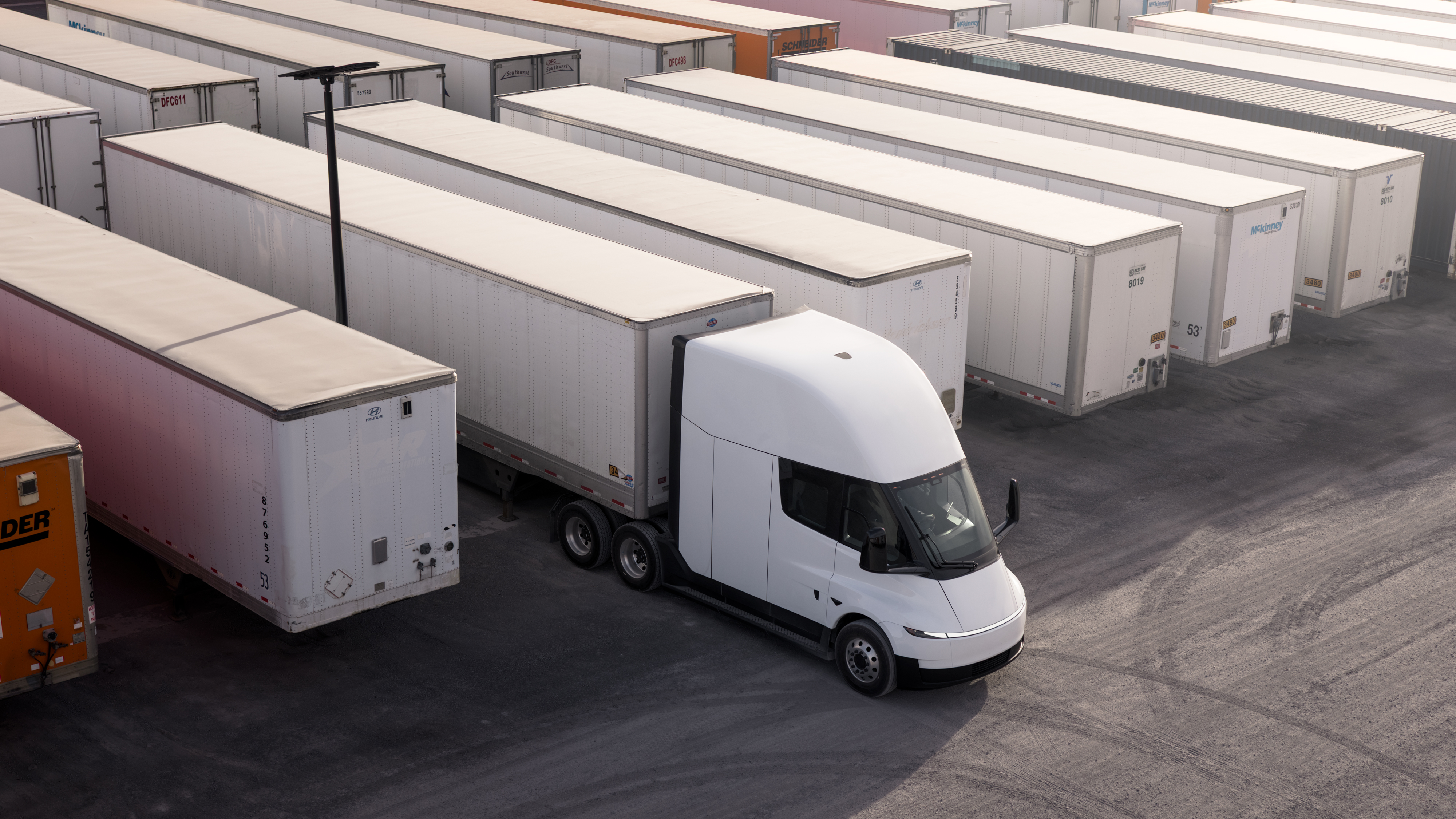 A Tesla Semi with a trailer is parked in a logistics yard alongside other trucks.