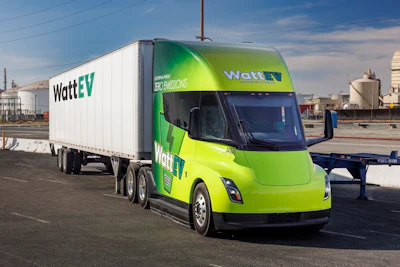 A green and yellow WattEV branded Tesla Semi and trailer parked at an industrial facility.