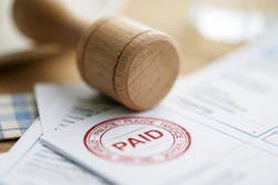 Wooden stamp creating a red 'PAID' mark on invoice documents at a desk